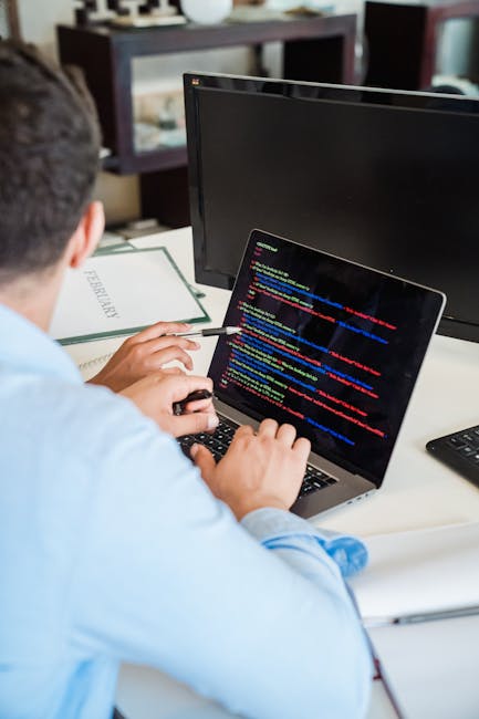 A person with dark hair, viewed from behind, is seated at a desk during a house removal process, working on a laptop displaying colorful lines of code, possibly related to technical setup or data tran