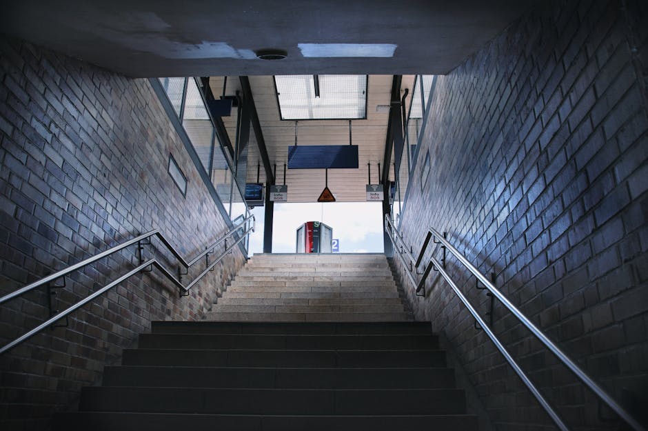 Inside view of a stairwell leading up from an underground station, with brick walls on either side and metal handrails along both sides of the staircase. The stairs are made of concrete or similar mat