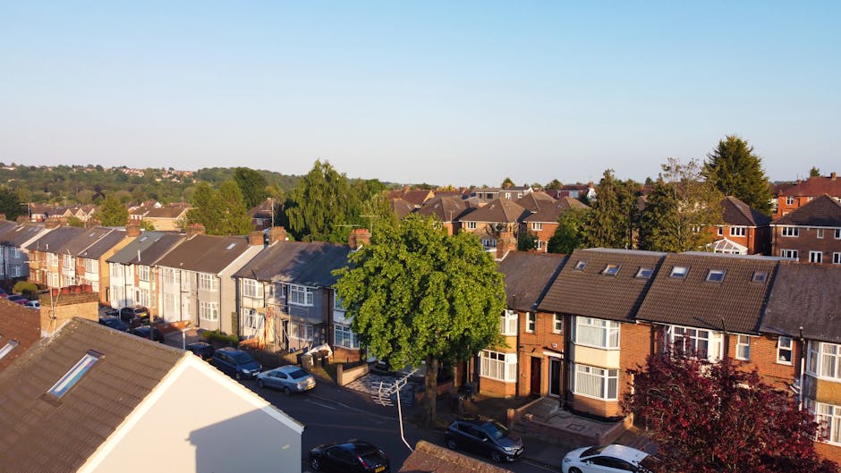 A residential street scene on a bright, sunny day with a clear blue sky, featuring a row of traditional terraced houses built with brick and light-colored stone, with multiple chimneys and large windo