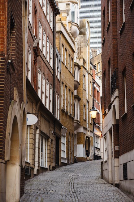 A narrow cobblestone alleyway situated between tall, multi-storey brick and stone residential buildings in an urban area. The buildings display a variety of architectural styles, with some featuring b